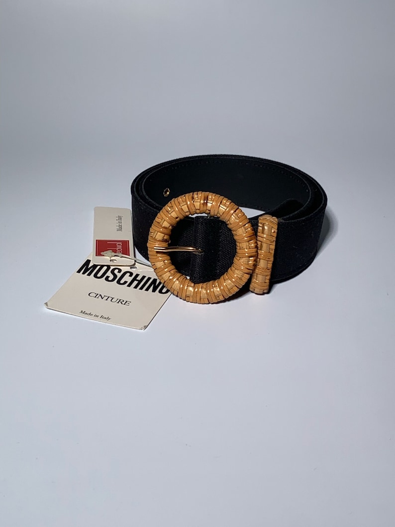 May include: A black woven belt with a large, round, woven buckle. The buckle is made of natural woven material and has a gold metal clasp. The tag attached to the belt reads "Moschino Cinture Made in Italy."