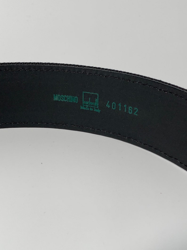 May include: Black leather belt with a green label that reads "Moschino" and "Made in Italy" with the number "401162" printed on the label.