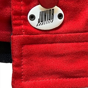 May include: Red jacket with a white oval patch that says "Junior Gaultier" in black lettering. The patch has two black buttons on either side. There is a red button above the patch.