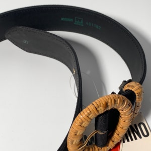 May include: A black canvas belt with a woven brown circle buckle. The buckle is made of natural materials and has a gold metal ring. The belt has a label that says "Made in Italy" and "Adventure".