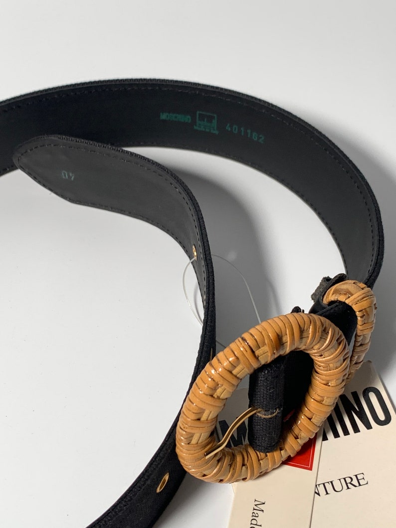 May include: A black canvas belt with a woven brown circle buckle. The buckle is made of natural materials and has a gold metal ring. The belt has a label that says "Made in Italy" and "Adventure".