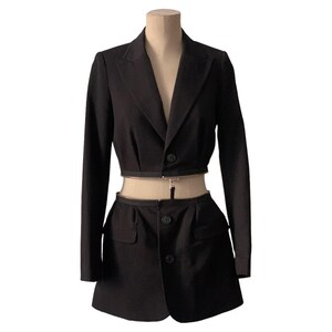 May include: A black cropped blazer and matching skirt set. The blazer has a single button closure and a zipper detail at the waist. The skirt has a single button closure and a pleated design.