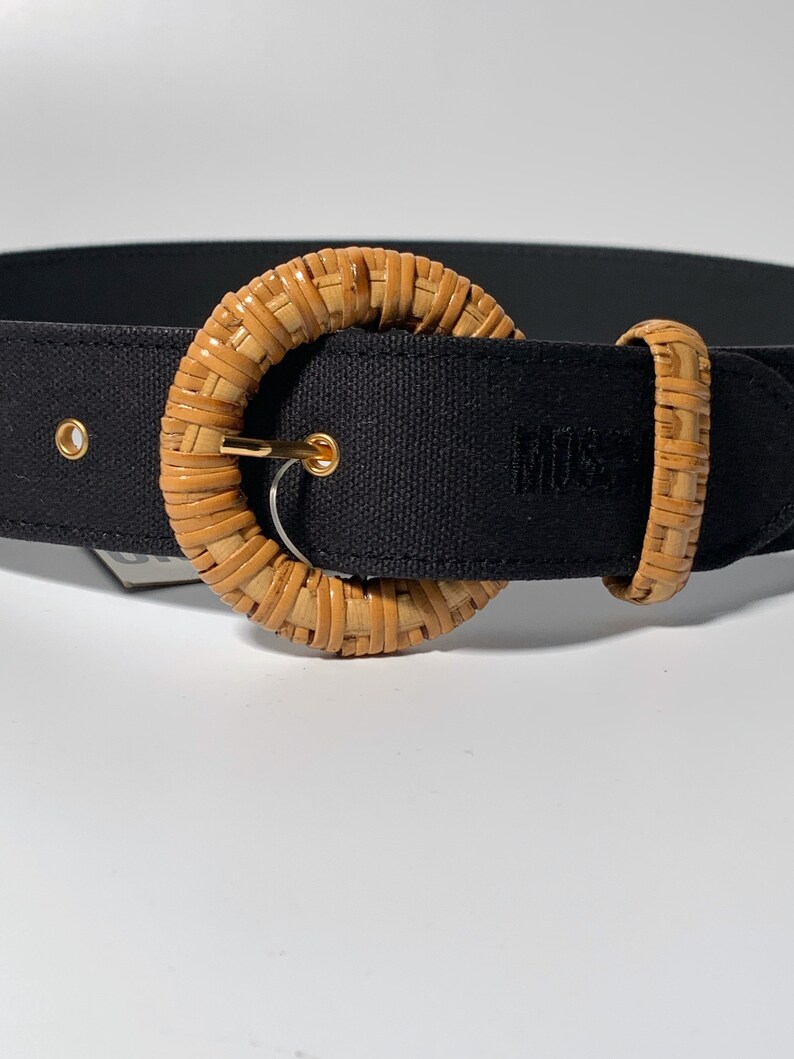 May include: A black canvas belt with a large, round, woven, light brown buckle. The buckle has a gold metal ring.