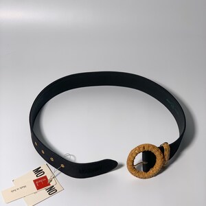 May include: Black leather belt with a woven, round buckle. The belt has gold-colored metal accents and is labeled "MONICA" and "Made in Italy".