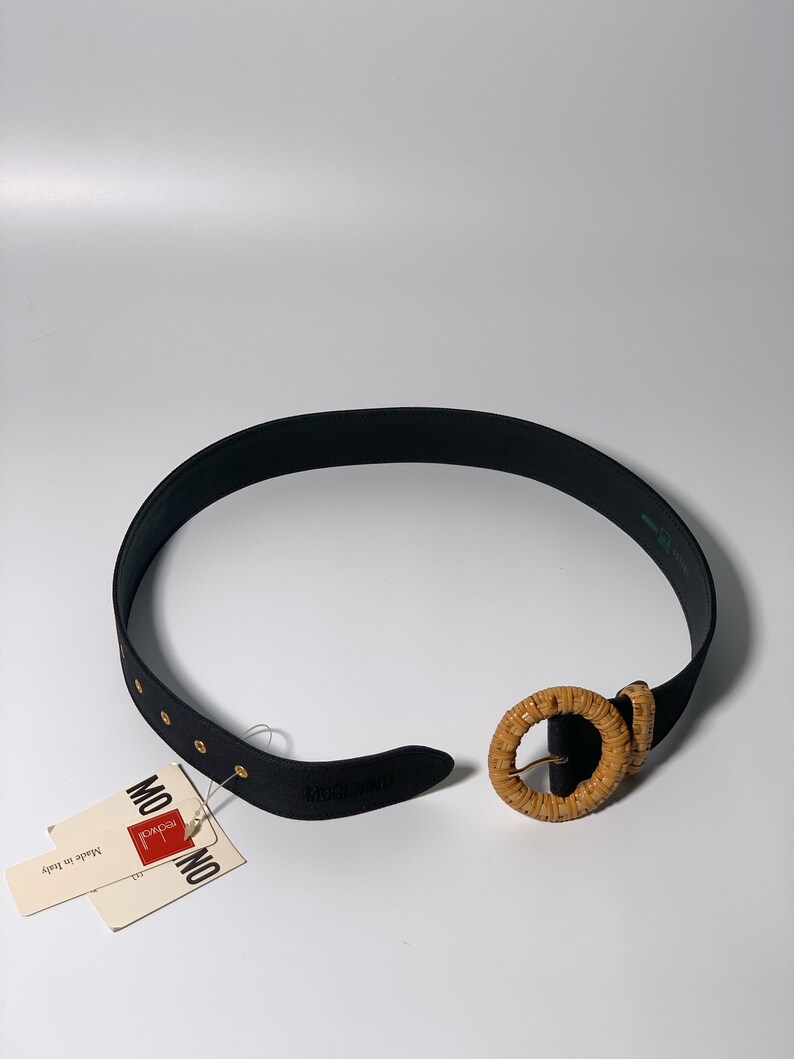 May include: Black leather belt with a woven, round buckle. The belt has gold-colored metal accents and is labeled "MONICA" and "Made in Italy".