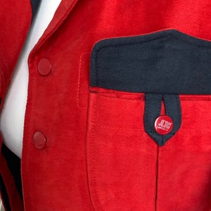 May include: Red jacket with a large black pocket on the front. The pocket has a smaller black pocket on the top with a red button that says "J.Crew" on it.