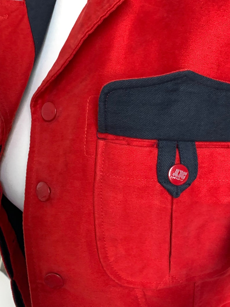 May include: Red jacket with a large black pocket on the front. The pocket has a smaller black pocket on the top with a red button that says "J.Crew" on it.