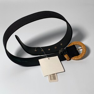 May include: A black fabric belt with a brown wooden buckle. The buckle is round and has a woven design. The belt has gold-colored metal accents.