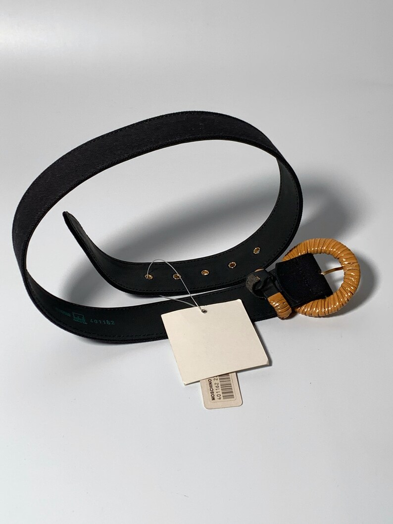 May include: A black fabric belt with a brown wooden buckle. The buckle is round and has a woven design. The belt has gold-colored metal accents.