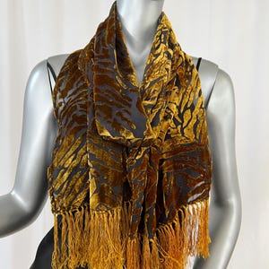 May include: A gold and black scarf with a velvet texture and fringe. The scarf has a pattern of gold stripes on a black background. The fringe is a matching gold color.