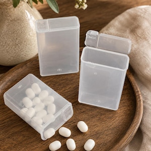 May include: Three rectangular, translucent plastic containers with hinged lids. One container is lying on its side with white, oval-shaped mints spilling out, while several loose mints are scattered nearby. The lids feature the embossed word OPEN.