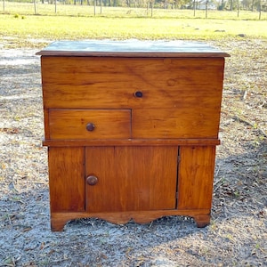 May include: A wooden cabinet with a single door and a drawer. The cabinet is a light brown color and has a simple design.