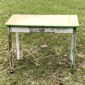 May include: A small, vintage table with a white base and a light green top. The table has a single drawer and is set on a grassy field. The paint is distressed, giving it a rustic appearance. The table is approximately 30 inches long.