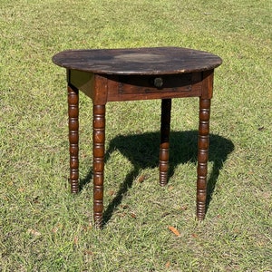 May include: A dark brown wooden table with a round top and a single drawer. The table has turned legs with a decorative design.