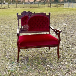 May include: A vintage red velvet armchair with carved wooden details sits in a grassy field against a backdrop of a cloudy sunset.