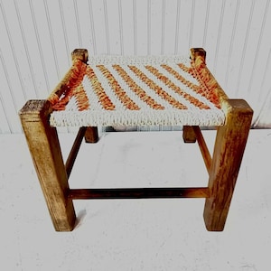 May include: A small, wooden stool with a woven seat. The seat is made of white and orange rope and has a striped pattern. The stool is sitting on a white surface.