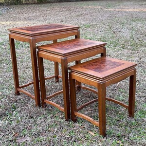 May include: Set of three nesting tables made of wood with a dark brown finish. The tops of the tables have a decorative inlaid pattern.