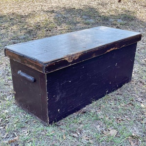 May include: A black wooden storage chest with a hinged lid and two metal handles. The chest is sitting on a grassy field with brown leaves scattered around it.