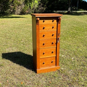 May include: A tall, wooden dresser with seven drawers and ornate carvings on the top. The dresser is a light brown color and has a slightly distressed finish. It is sitting on a grassy lawn.