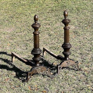 May include: A pair of antique cast iron fireplace andirons with ornate details. The andirons are brown and have a decorative design on the base.