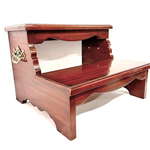 May include: A two-step wooden step stool with a rich, reddish-brown finish. The stool features a decorative brass handle on the side and scalloped edges. The top step is approximately 12 inches wide, and the bottom step is about 18 inches wide.