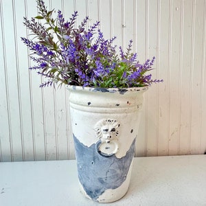 May include: A white ceramic vase with a distressed finish and a lion head design. The vase is filled with purple lavender flowers.