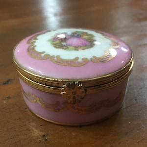 May include: A small pink and gold trinket box with a floral design and a couple in a romantic scene on the lid.