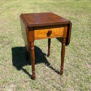 May include: A small, antique wooden side table with a dark brown top and a single drawer. The drawer has a round wooden knob. The table has four slender, turned legs. The table's dimensions are approximately 24 inches high, 20 inches wide, and 20 inches deep.