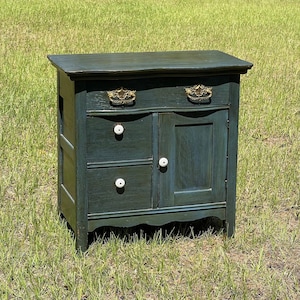 May include: A dark green painted wooden cabinet with two drawers and a door. The cabinet has ornate gold handles and white knobs. The cabinet is sitting on a grassy lawn.