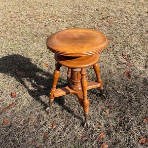 May include: A wooden stool with a round seat and four legs. The legs are turned and have a decorative design. The stool is in a grassy area.