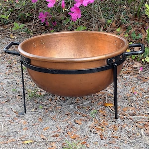 May include: A large copper cauldron with black iron legs. The cauldron is sitting on a bed of brown leaves and pine needles.