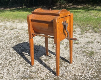 May include: Antique wooden butter churn with a hand crank. The churn is brown and has a wooden lid. It sits on four legs and is in good condition.