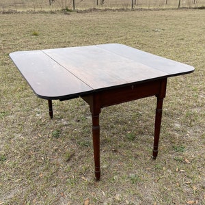 May include: A dark brown, antique drop-leaf table with a rectangular top and rounded corners. The table has a drawer and four turned legs. The table is set up outdoors on a grassy field.