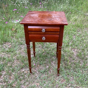 May include: A small wooden end table with two drawers and turned legs. The table has a dark brown finish and is sitting on a bed of green grass.