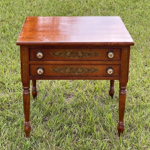 May include: A wooden end table with two drawers and turned legs. The drawers have gold painted floral designs and brass knobs. The table is sitting on a green grassy lawn.