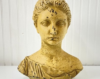 May include: A vintage-style bust of a woman with a classical design. The sculpture is painted in a muted yellow color with dark accents, giving it an aged appearance. The bust features detailed facial features and a draped garment.