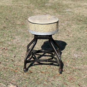 May include: An antique adjustable stool with a round, patterned seat and a black metal base. The seat has a beige and yellow woven design with a silver metal band. The base features a decorative, wrought-iron style design.