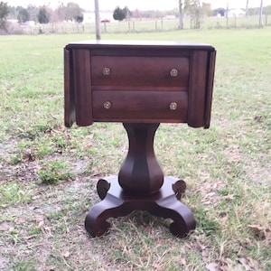 May include: A dark brown wooden end table with two drawers and a pedestal base. The table has a rounded top and decorative feet.