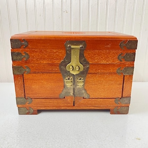 May include: A wooden jewelry box with a reddish-brown finish. The box features brass hardware, including a central clasp and corner accents. The box has a rectangular shape with a hinged lid and two small doors. The box is elevated on small feet.