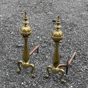 May include: A pair of antique brass fireplace andirons with ornate finials and three legs. The andirons are on a gravel surface.
