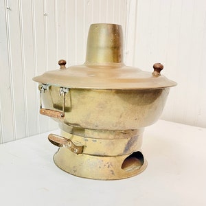 May include: A vintage brass hot pot with wooden handles. The pot has a round base with a hole in the center and a tall, cylindrical top. The pot is designed for cooking over a small flame.