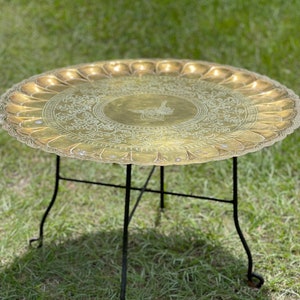 May include: A round brass tray with an intricate floral design and a black metal stand. The tray has a scalloped edge and is perfect for serving food or drinks.