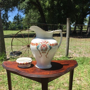 May include: A white ceramic pitcher with a floral design sits on a dark wood table with a matching ceramic bowl. The table has a curved top and three legs.