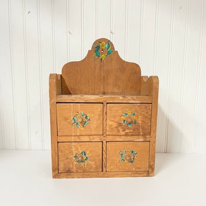 May include: A wooden wall-mounted storage unit with four drawers. Each drawer has a painted floral design with yellow flowers and green leaves. The top of the unit has a decorative arched top with a similar floral design.