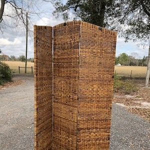 May include: A three-panel room divider made of woven wicker. The panels are brown and have a natural finish. The divider is standing on a gravel driveway.