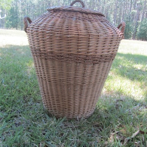 May include: A large, brown wicker basket with a lid and two handles. The basket is sitting on a bed of green grass.