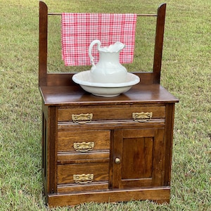 May include: A vintage wooden washstand with a white porcelain pitcher and bowl. The washstand has three drawers and a cabinet door with brass hardware. A red and white checkered towel hangs on the back.