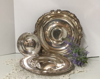 May include: Three silver serving dishes of varying sizes, with ornate edges, arranged on a white crocheted doily.