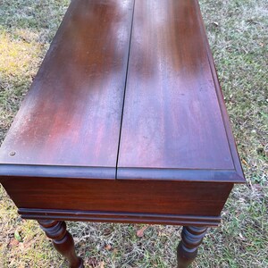 May include: A dark brown wooden table with a hinged top and turned legs. The table is sitting on a bed of green grass.