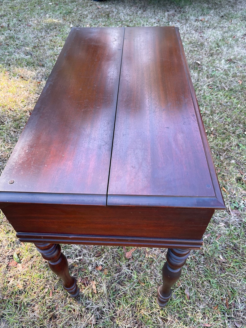 May include: A dark brown wooden table with a hinged top and turned legs. The table is sitting on a bed of green grass.